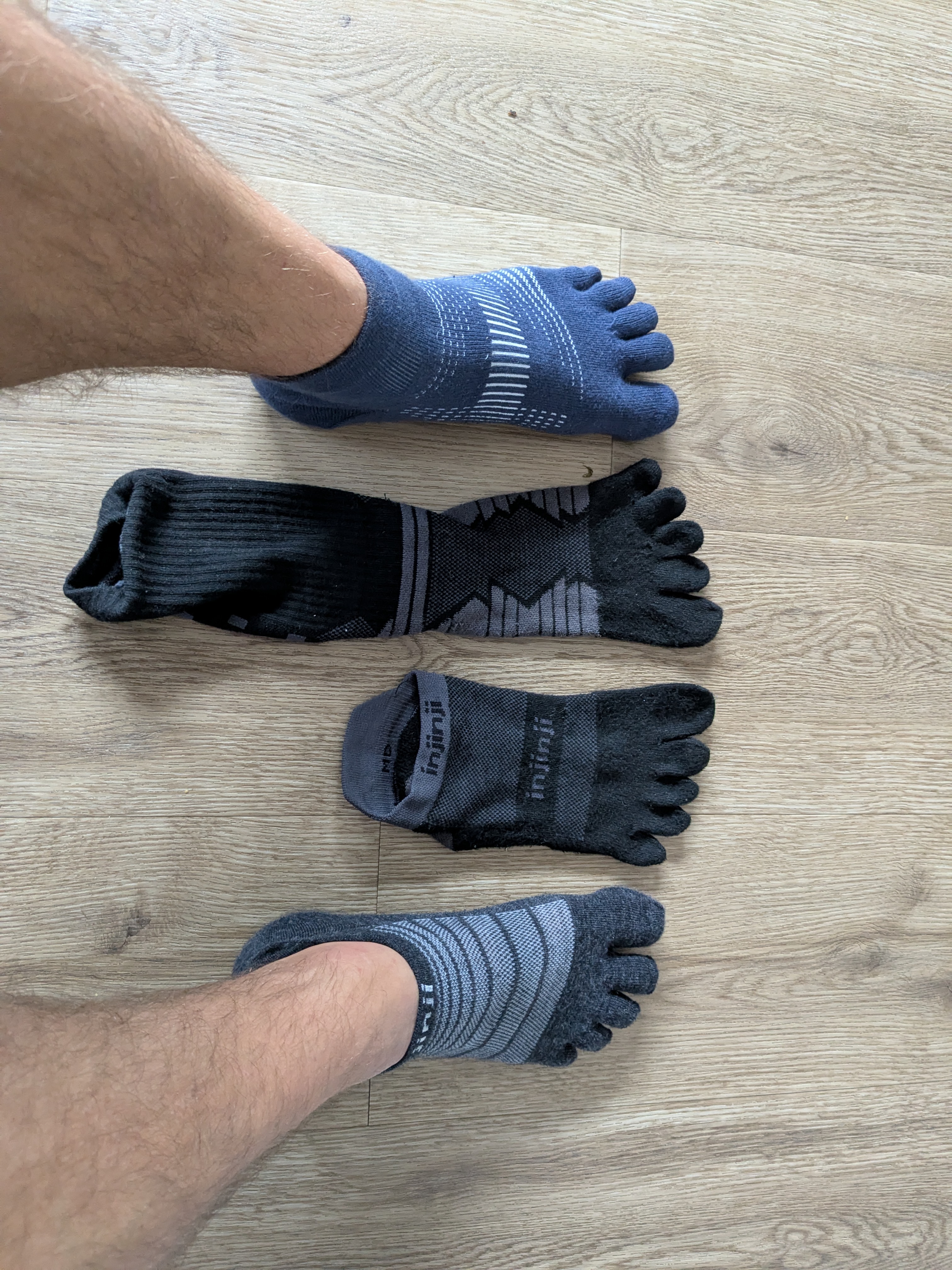Four Injinji toe sock variants laid out side by side — blue mini-crew, black crew, grey merino no-show, and black midweight no-show